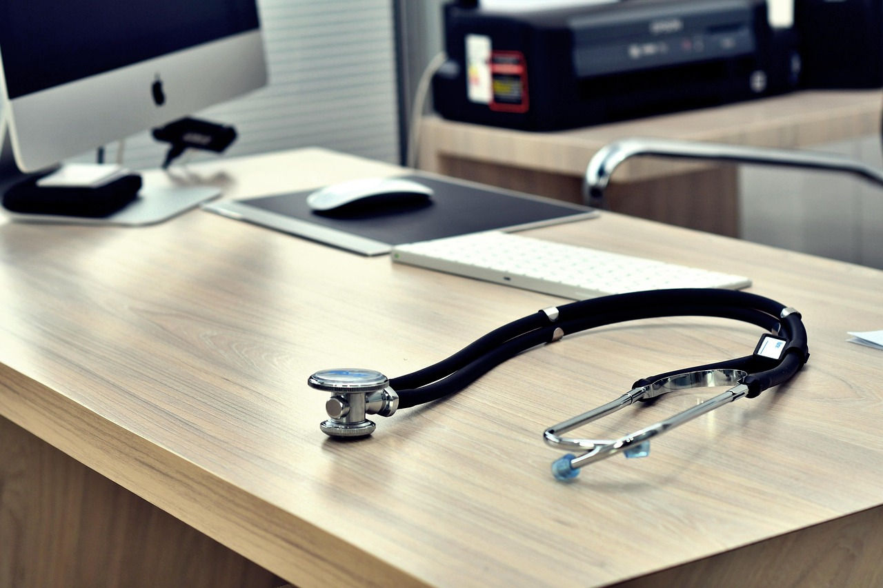 Stethoscope on a wooden desk with an iMac, keyboard, and printer in a modern, organized office setting. Calm and professional mood. - doctors offer same day appointments
