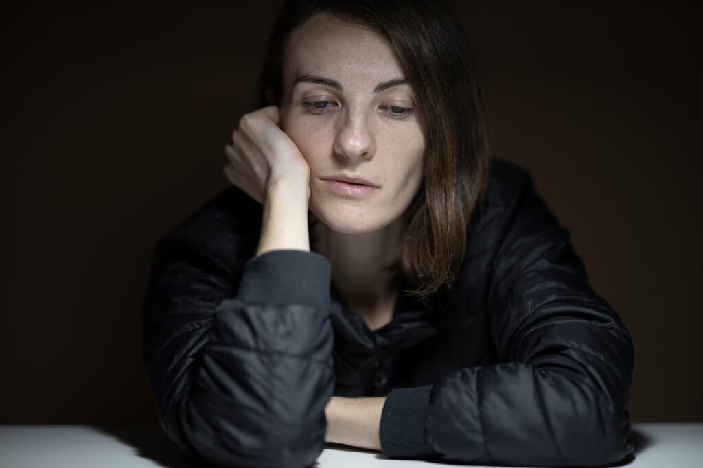 A woman wearing a black jacket rests her head on her hand, looking down with a tired and pensive expression in a dimly lit room- Burnout vs. Depression