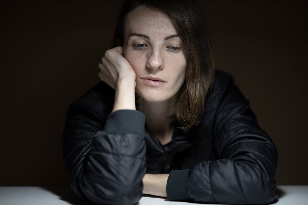 A woman wearing a black jacket rests her head on her hand, looking down with a tired and pensive expression in a dimly lit room- Burnout vs. Depression