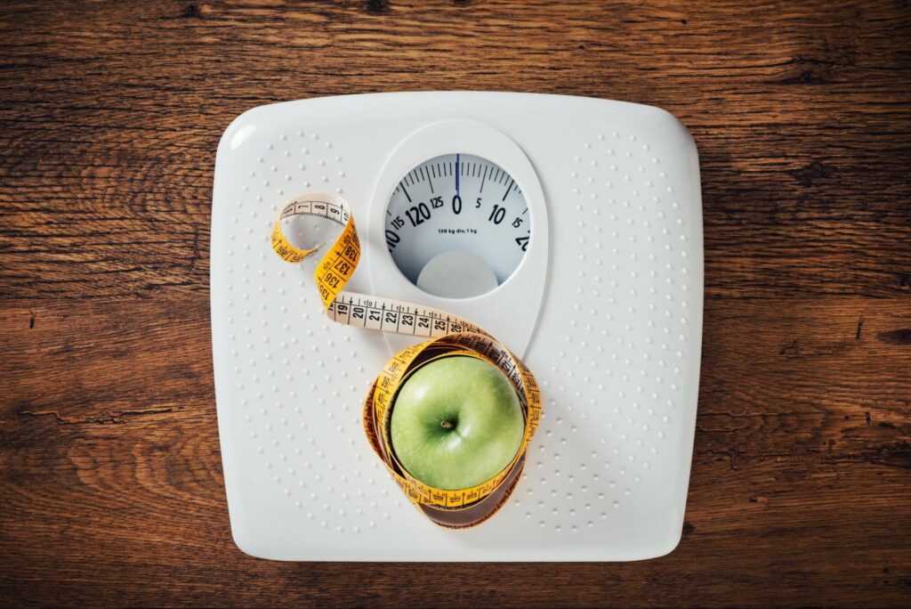 apple wrapped with tape measure on weighing scale, representing the need for weight management and the benefits of having a weight loss doctor in Houston TX