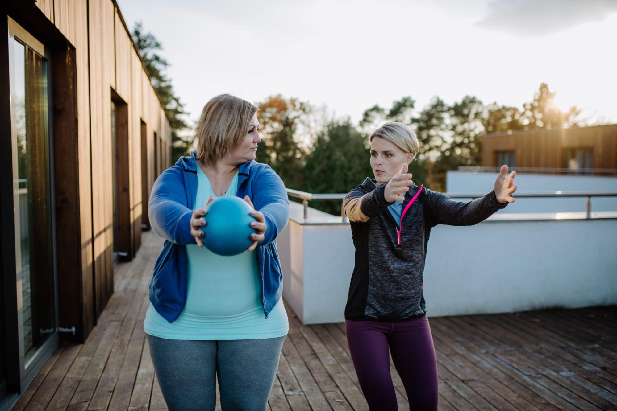 Overweight woman holding ball and exercising with personal trainer in outdoors on gym terrace